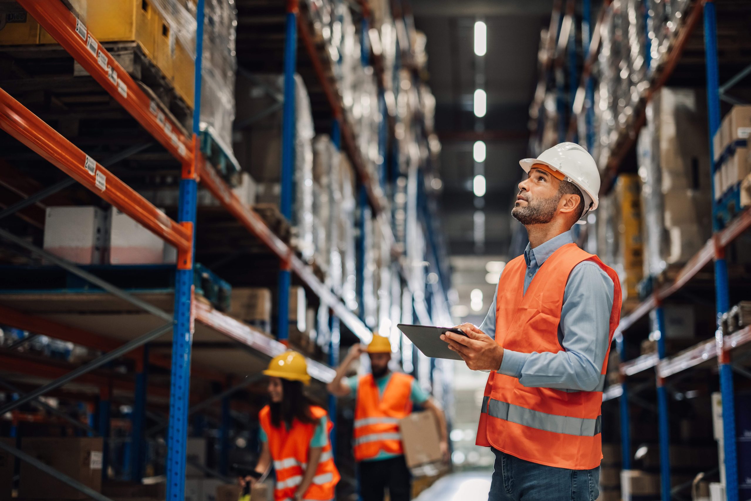 Warehouse manager holding digital tablet, looking up at shelves while checking inventory with his team in the background