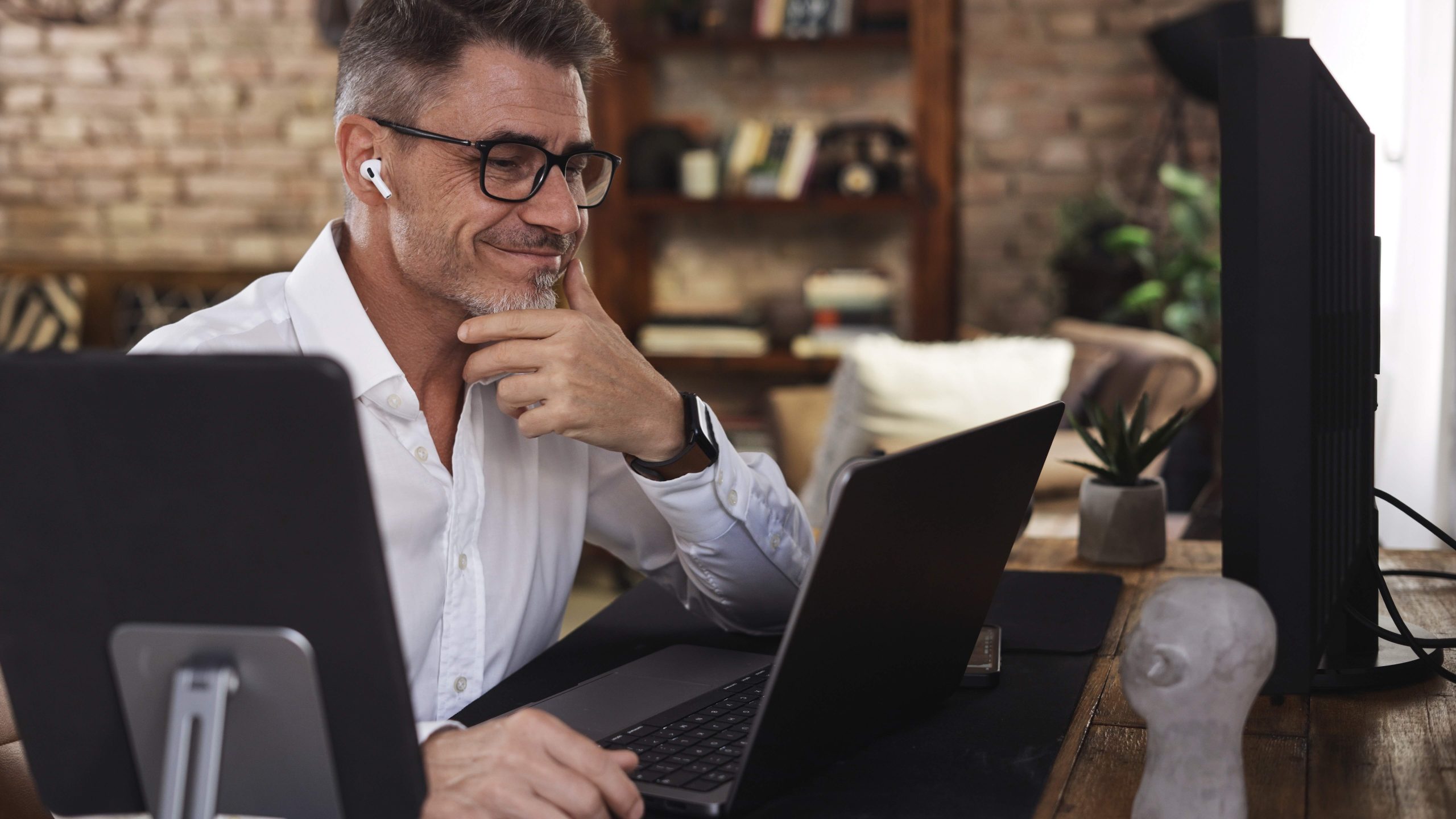 Businessman working from home office typing on laptop using digital tablet. Happy mid adult man in glasses sitting at desk, smiling, thinking. Successful male entrepreneur leading business online.