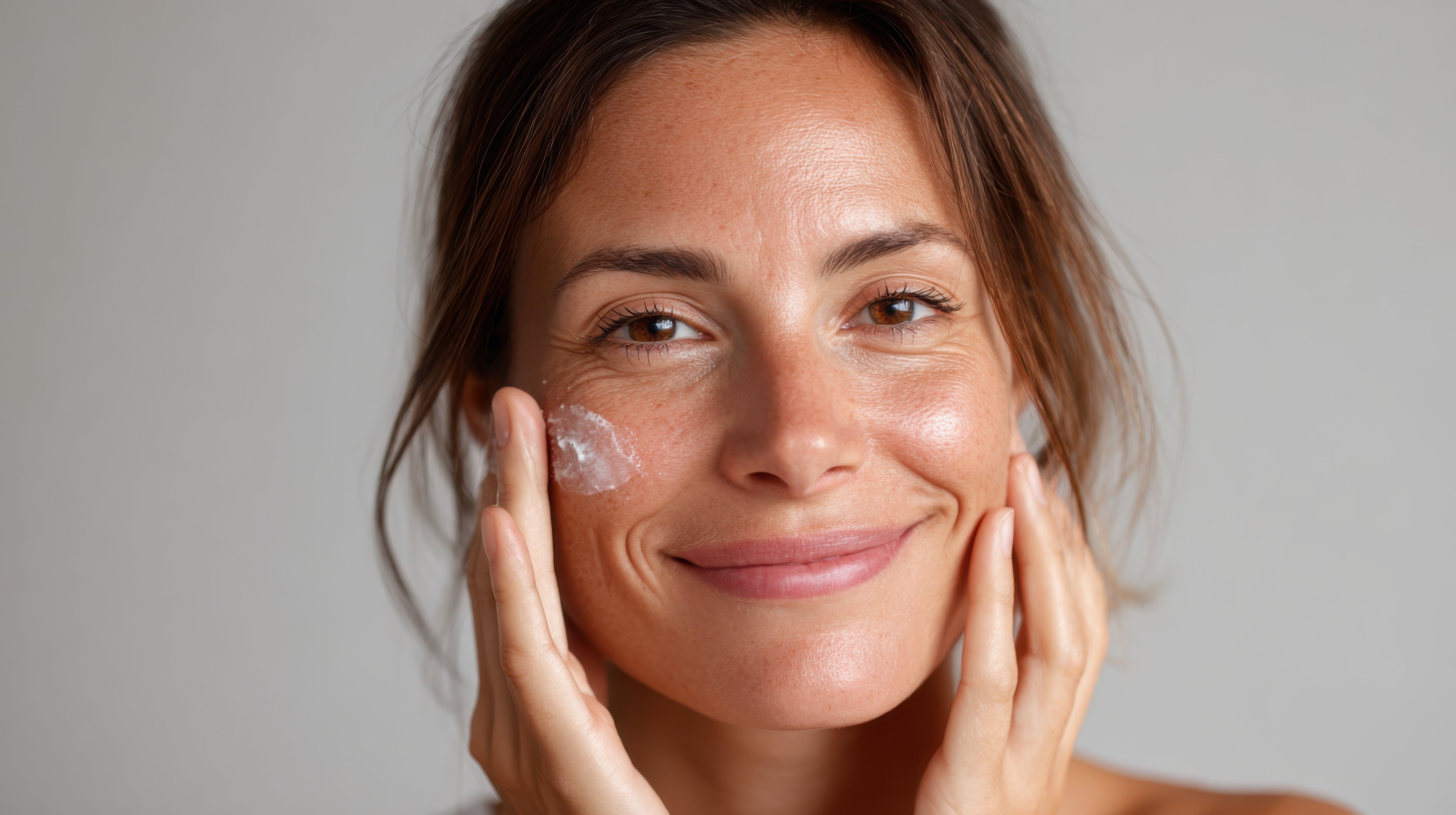 Smiling woman applying moisturizer cream to her cheek, showcasing healthy, radiant skin with visible fine lines and freckles.