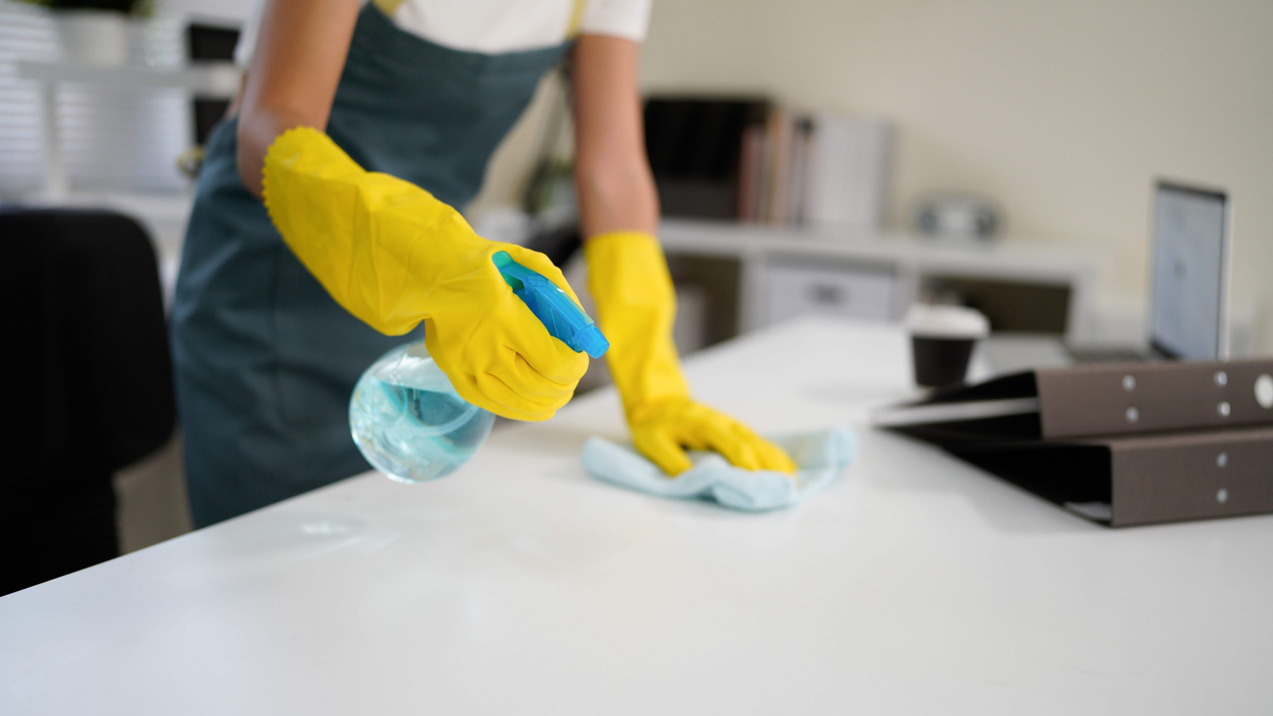 Young woman wearing yellow gloves and an apron, smiling while cleaning a white table with disinfectant spray and a blue cloth, promoting hygiene and cleanliness in her workspace.