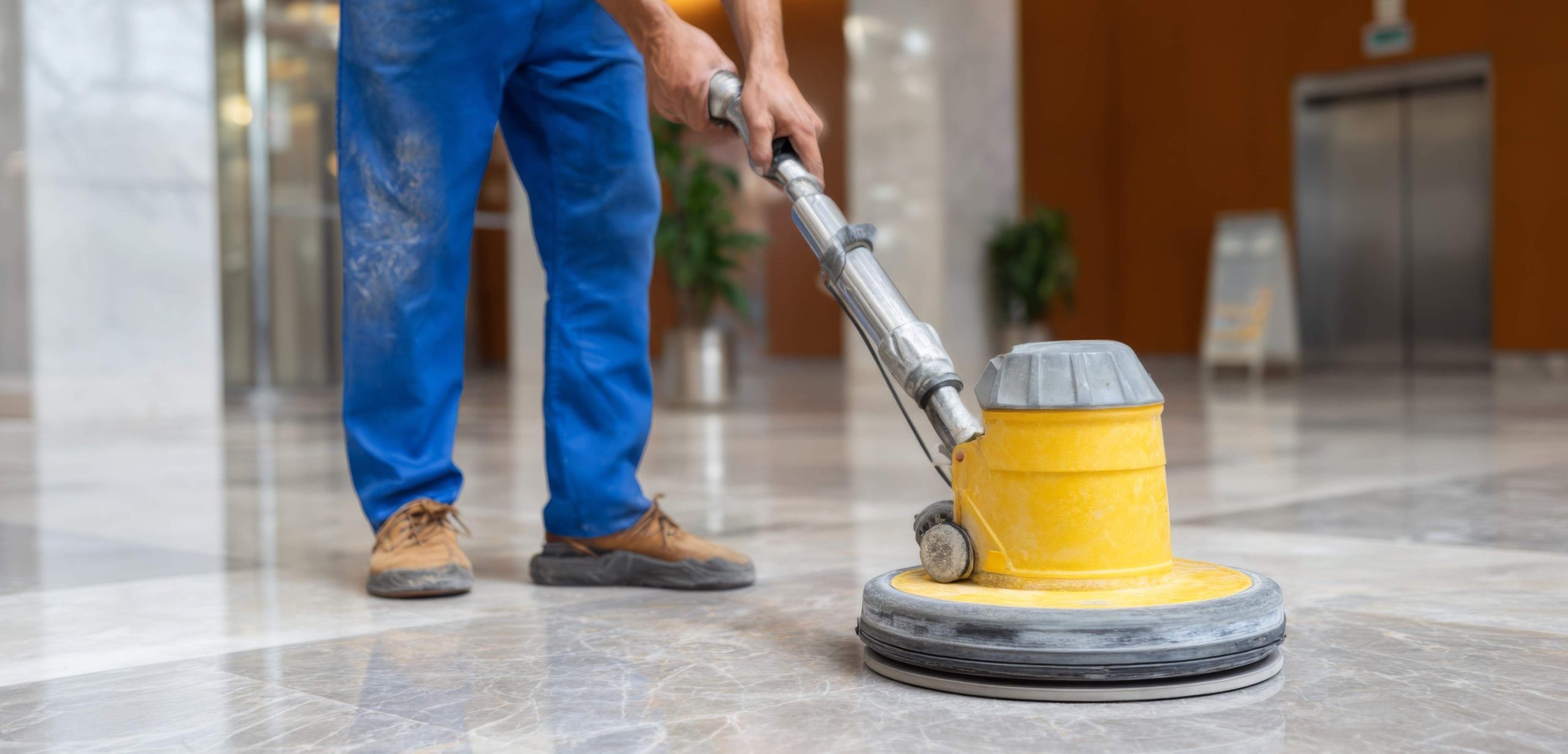 A laborer in blue overalls cleans a marble floor with a yellow rotary floor machine, showcasing diligent work in a well-lit environment.