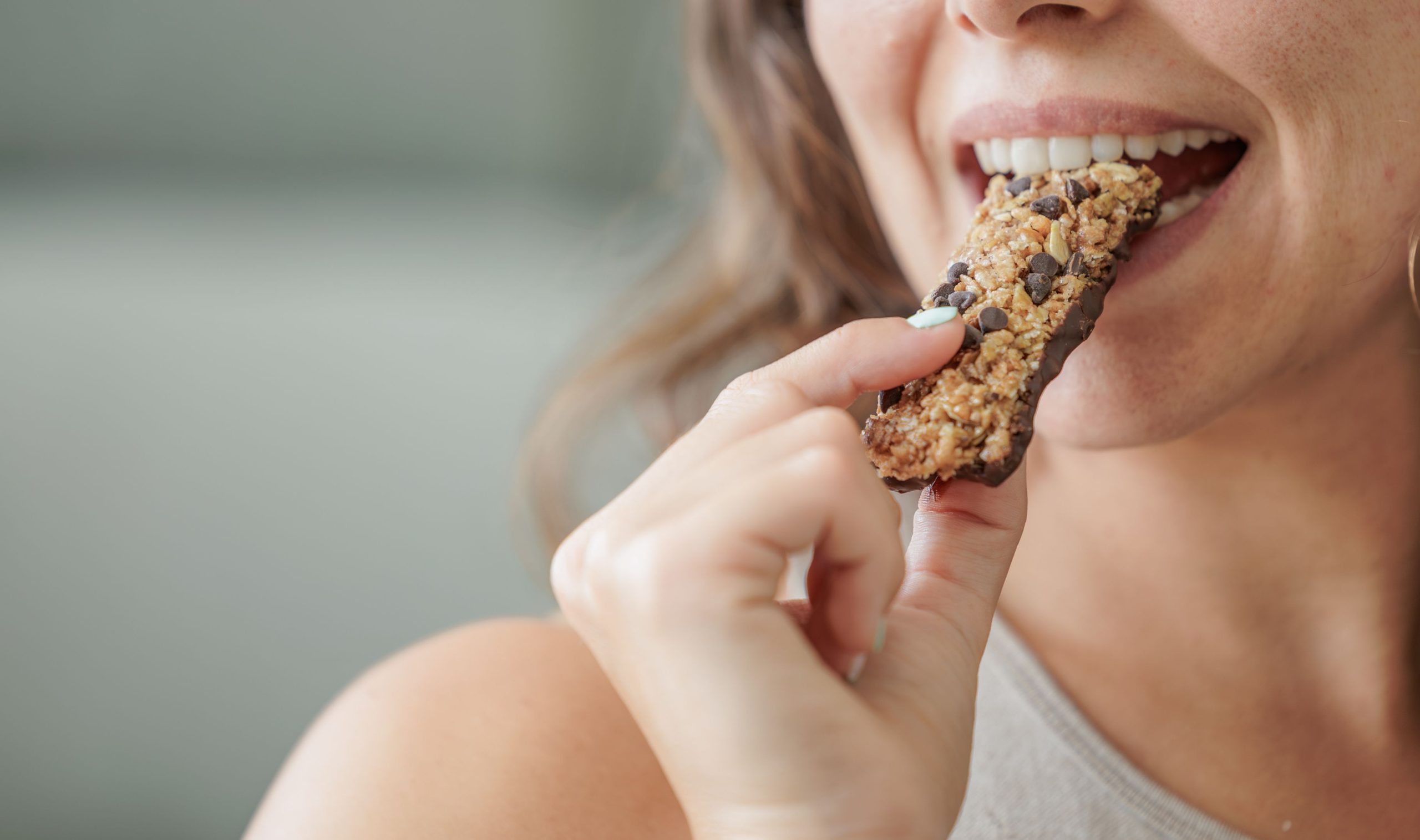 Portrait of a smiling young woman eating a protein bar with chocolate chips.